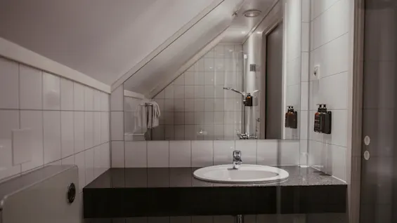 Et lyst og ryddeligt hotelbadeværelse med håndvask, spejl og bruseområde, med hvide fliser og skråloft.