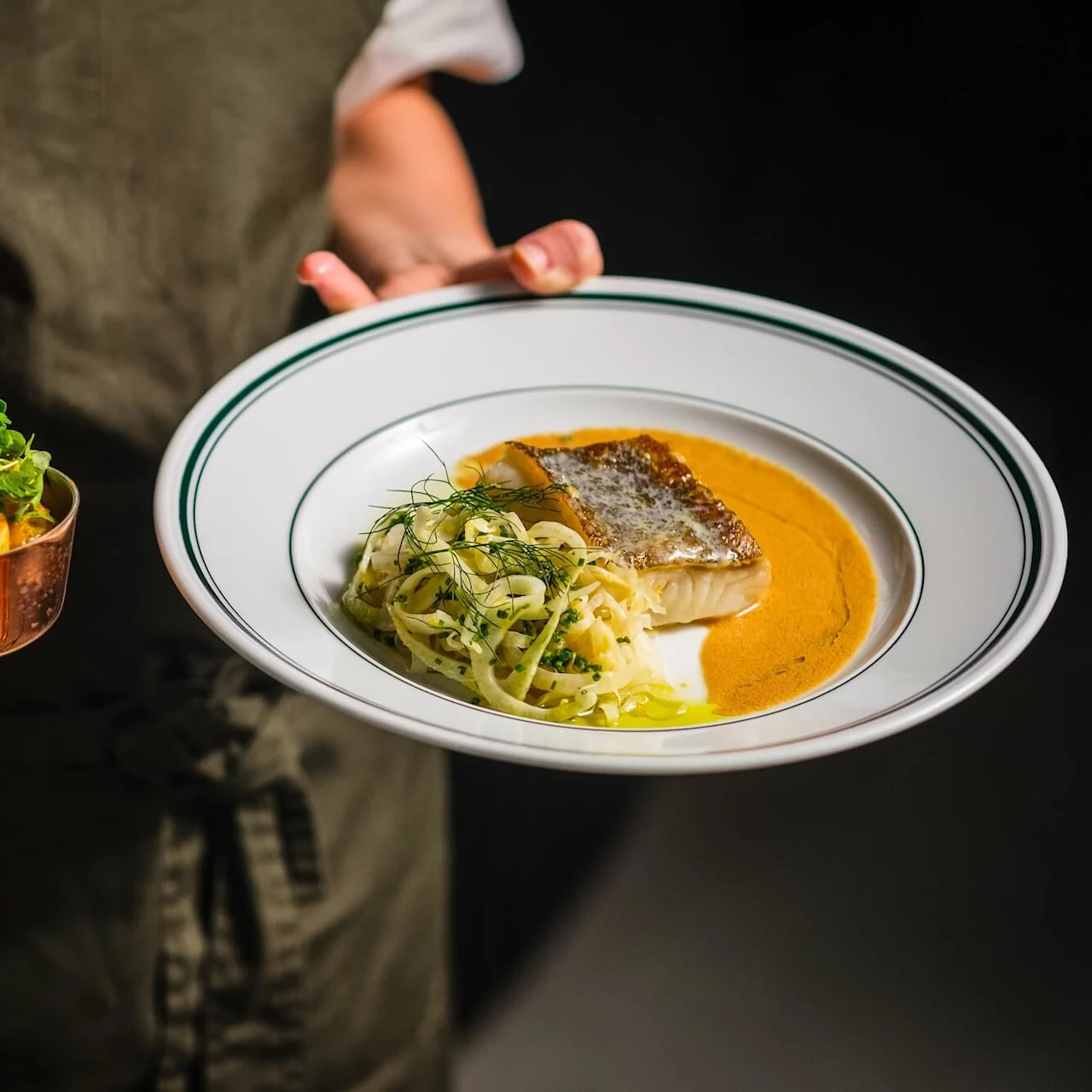En tallrik med brynt fisk, pasta och sås hålls av en person i ena handen, medan den andra håller en liten kopparkastrull med garnityr. Personen bär ett mörkt förkläde.