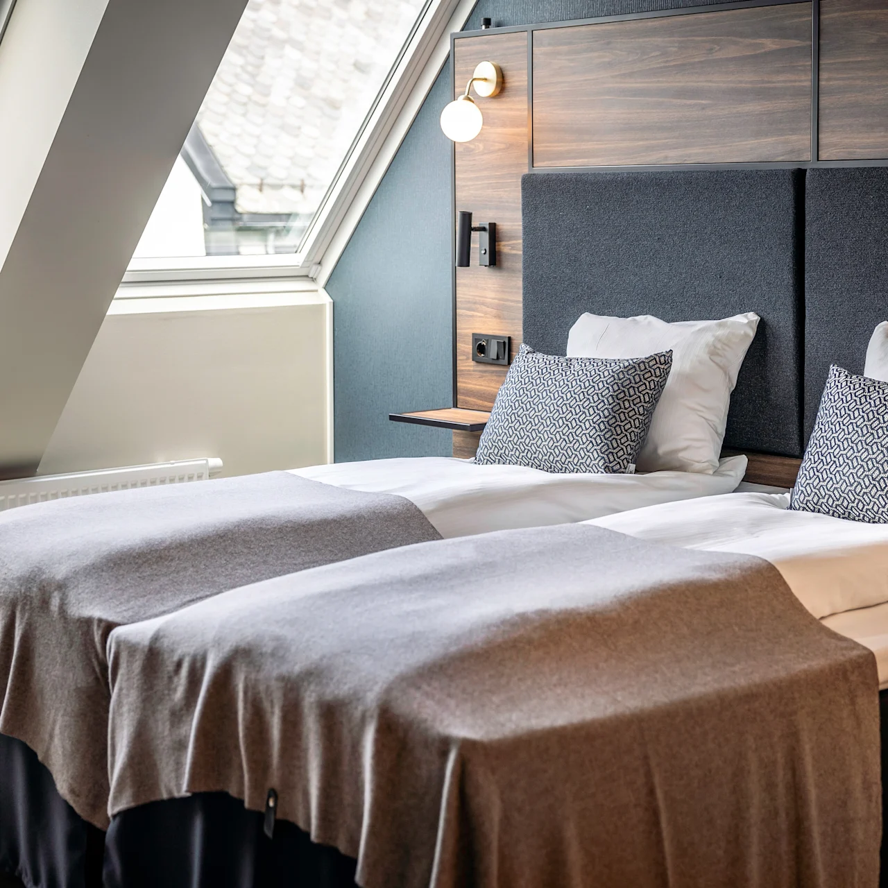 Två prydligt bäddade enkelsängar med gråa filtar och mönstrade kuddar står i ett modernt rum. Stora fönster ger naturligt ljus, med en träpanel och lampa ovanför sängen.