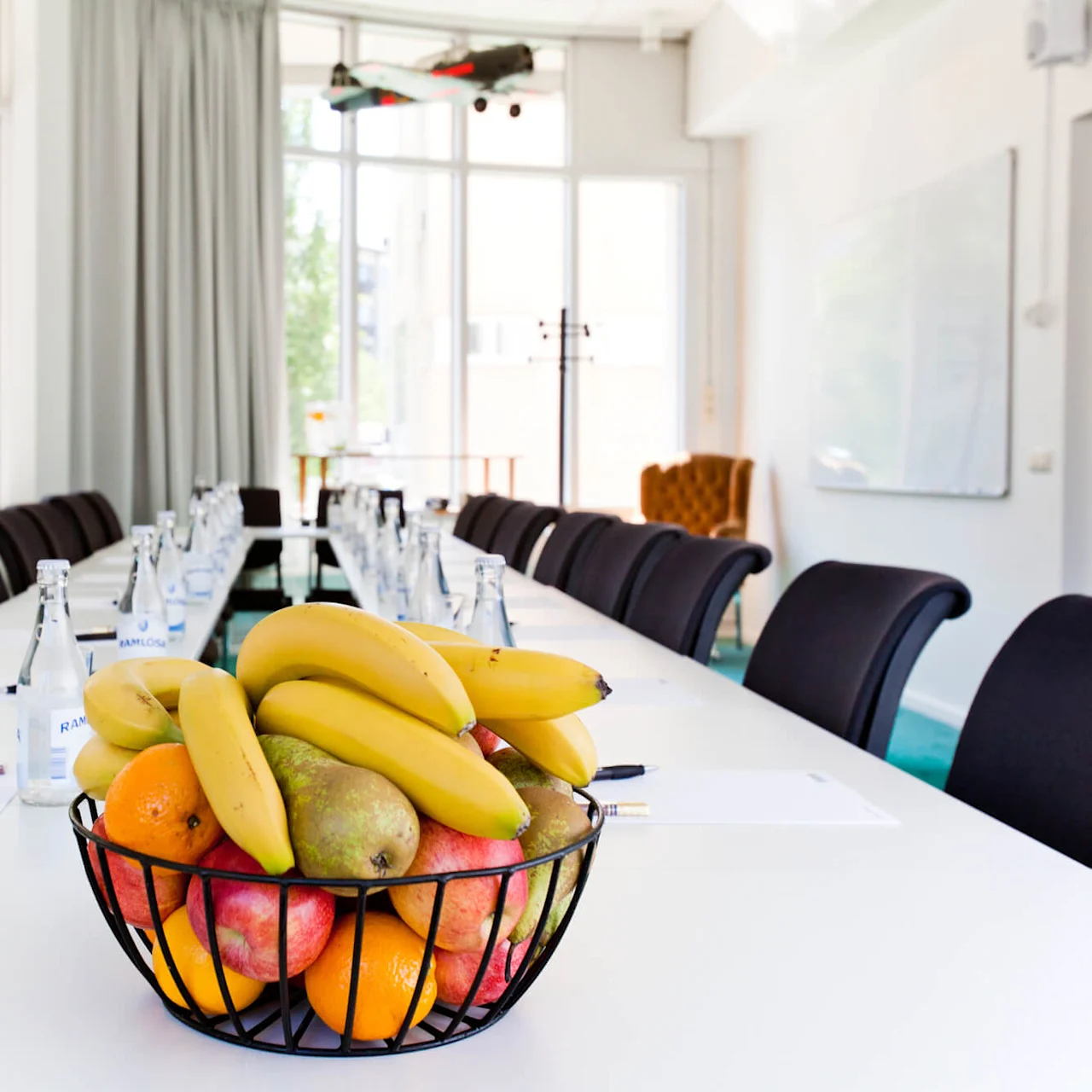 A fruit basket on a conference table with water bottles, glasses, and notepads; bright room with large windows.