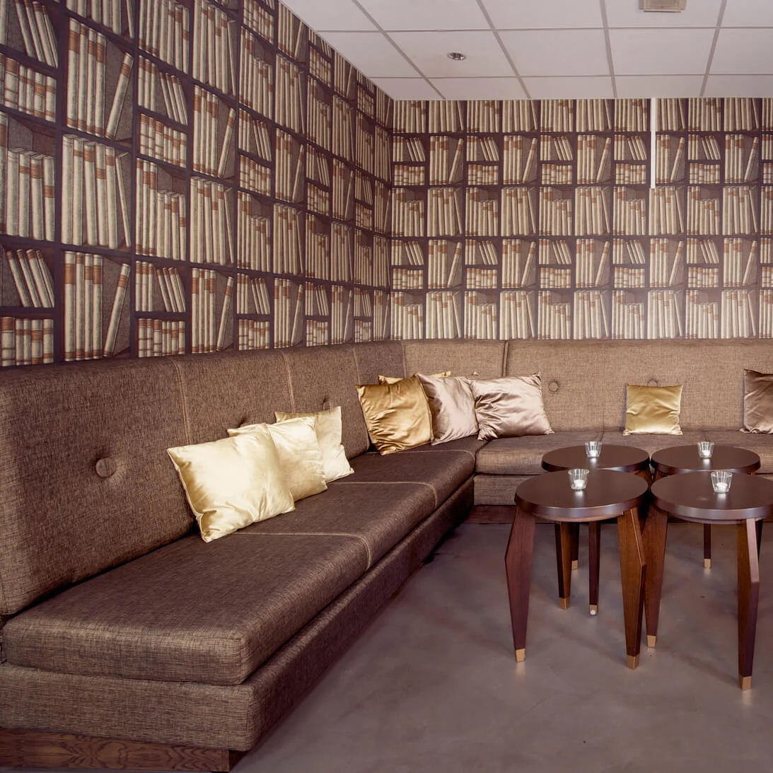 A corner sofa with gold and brown pillows, round coffee tables in front, against a wallpaper mimicking a bookshelf.