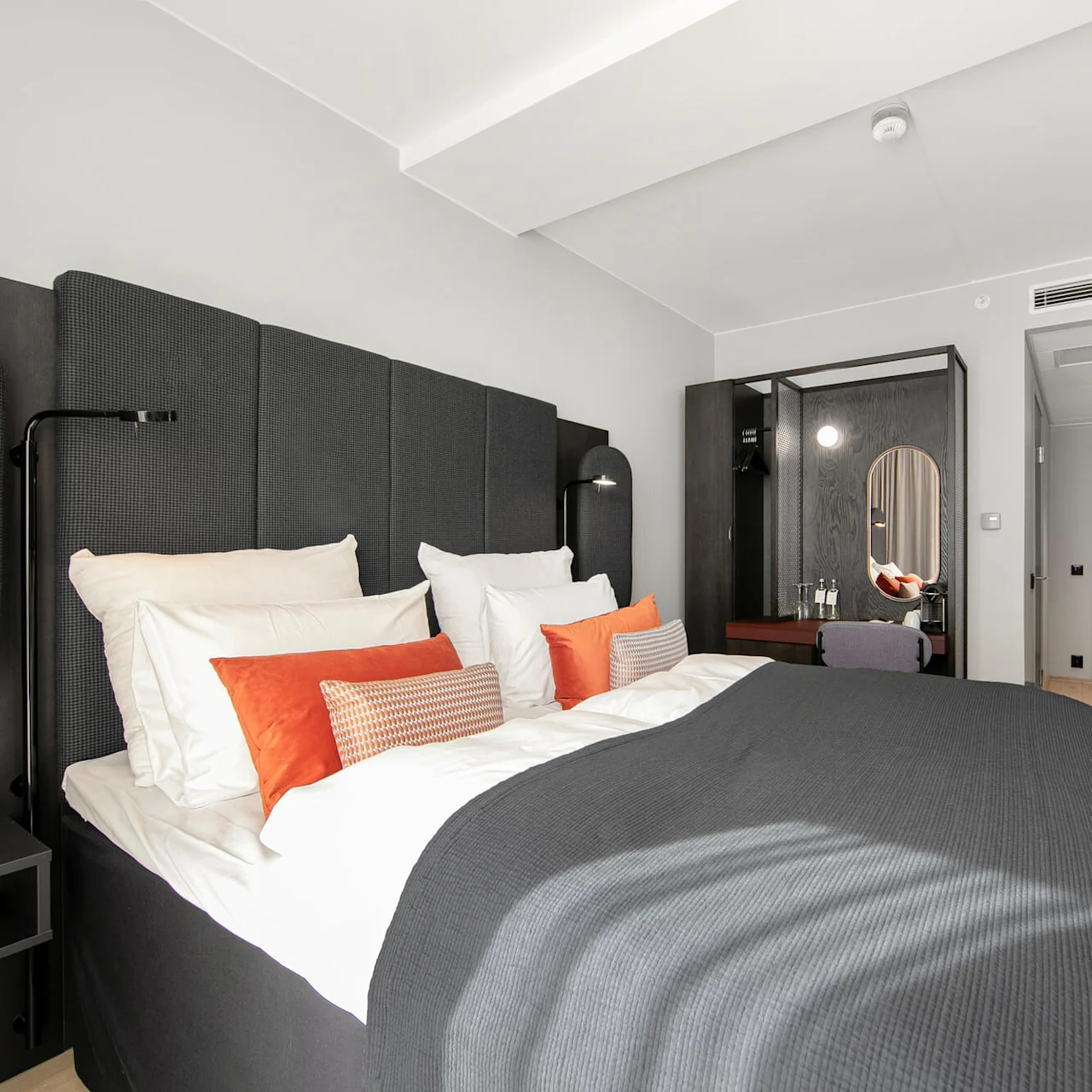 A neatly made bed with gray and orange pillows sits in a modern hotel room. A mirror, desk, and door are in the background.