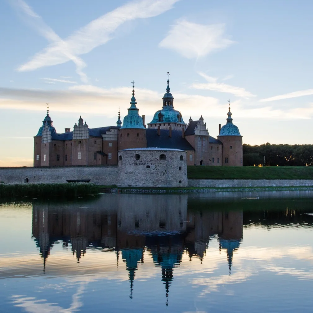Et storslået slot med flere tårne og kupler spejlet i roligt vand under en blå himmel med skyer.