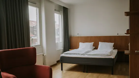 En pænt redt dobbeltseng med hvidt sengetøj står op mod en træpanel i et rum med lyse trægulve, en rød lænestol og store vinduer med grå gardiner.