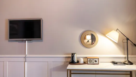A hotel room with a wall-mounted TV, a desk with a lamp, a mirror, and coffee-making facilities, creating a cozy and functional space.