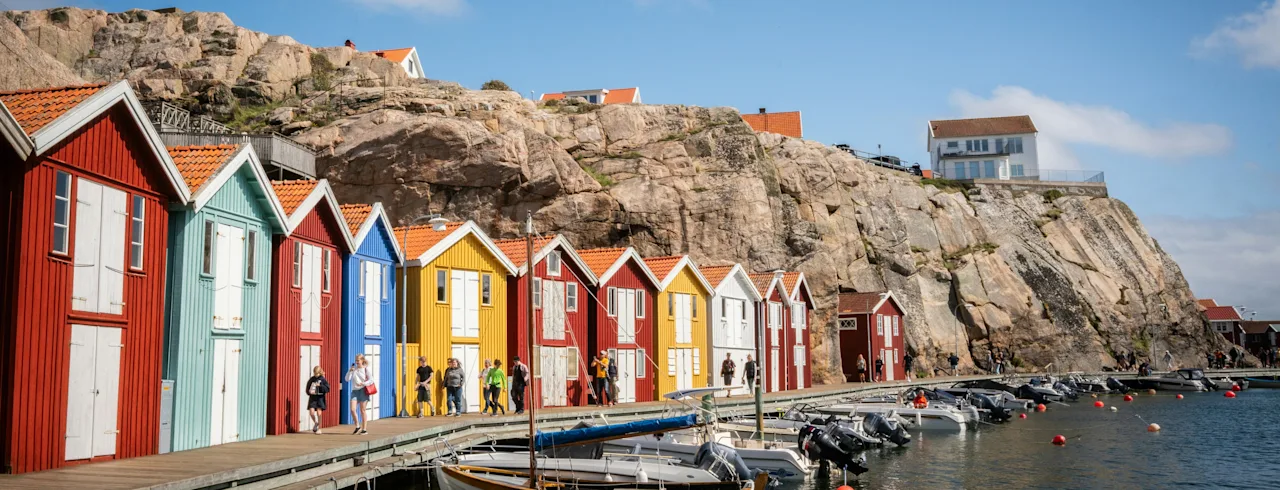 Colorful boathouses line a waterfront with boats docked, people walking, and a rocky cliff in the background.