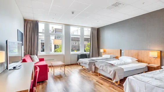 Hotellihuone, jossa on kaksi sänkyä valkoisilla vuodevaatteilla ja seinälamput puulattialla. Punainen sohva ja työpöytä, jossa TV, ovat suurien ikkunoiden lähellä.