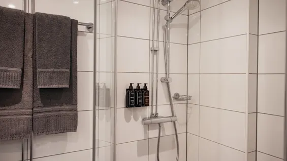 En ren, modern dusch med vita kakelplattor, ett duschhuvud, toalettartiklar och fräscha handdukar redo att användas.