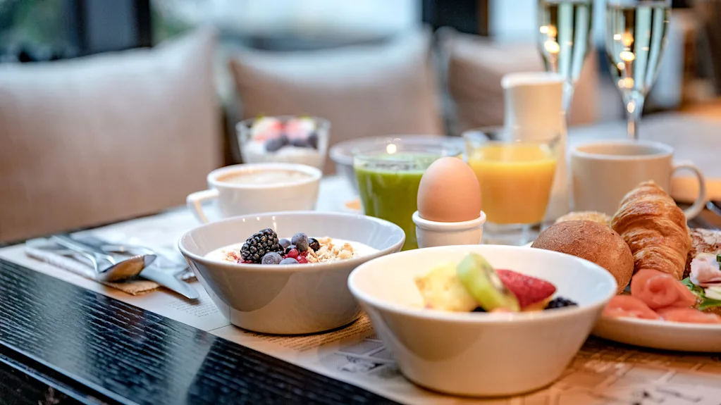 En frukost bestående av skålar med frukt och yoghurt, ett löskokt ägg, croissant, kaffe, juice och champagne, på ett träbord i en mysig inomhusmiljö.