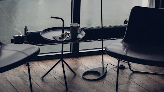 A round black table holds a coffee mug, chocolate, and spoon, situated between two chairs on wooden flooring by a window, creating a cozy atmosphere.