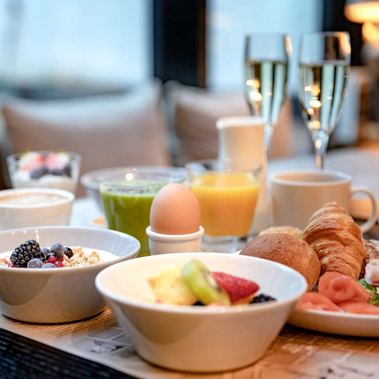 En frokost består av skåler med frukt og yoghurt, et bløtkokt egg, croissant, kaffe, juice og champagne, på et trebord i en koselig innemiljø.
