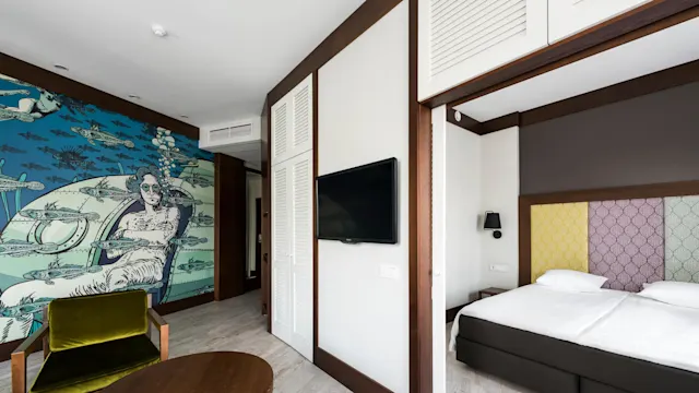 A room with a green chair and round wooden table beside a bed. A mural of a woman in a submarine with fish adorns the wall. A TV is mounted above a cabinet.