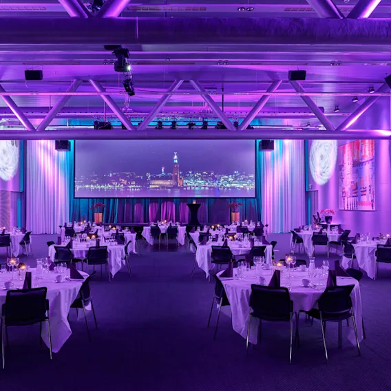 A spacious banquet hall features round tables with white tablecloths, elegantly set with glasses and cutlery. Purple lighting creates a vibrant atmosphere, and a large screen displays a city skyline.