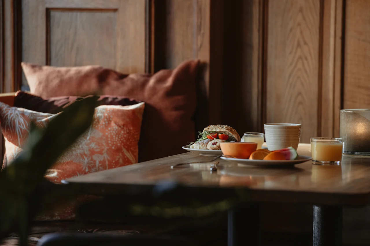 En mysig frukost med en smörgås, färsk frukt och juice på ett träbord, med mjuka kuddar i bakgrunden. Home Hotel Grand, Helsingborg.