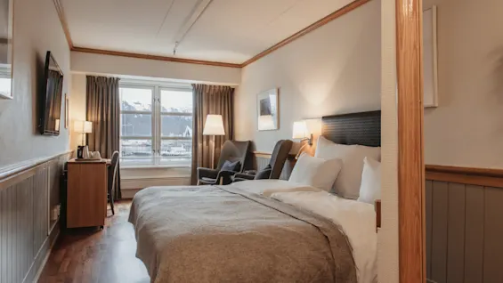 A cozy hotel room with a comfortable bed, a desk, and armchairs by a window overlooking snowy mountains.