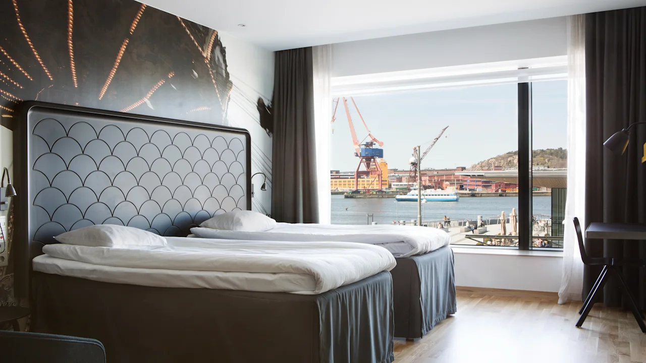 Ett hotellrum med två sängar och ett stort fönster som erbjuder utsikt över en livlig hamn med kranar och vatten.