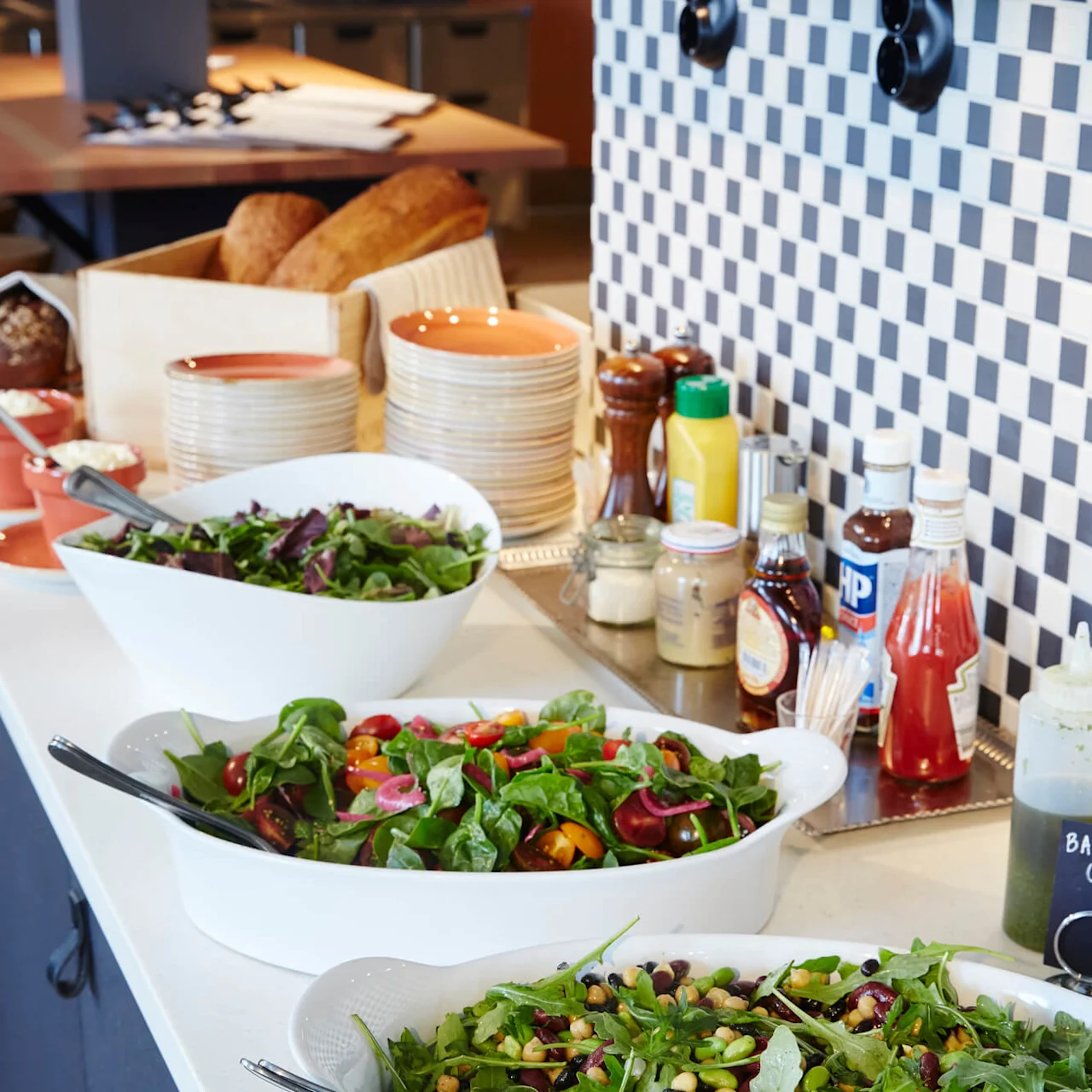 Fresh salads in white bowls are displayed on a counter for self-service, accompanied by bread and condiments, in a casual dining setting.