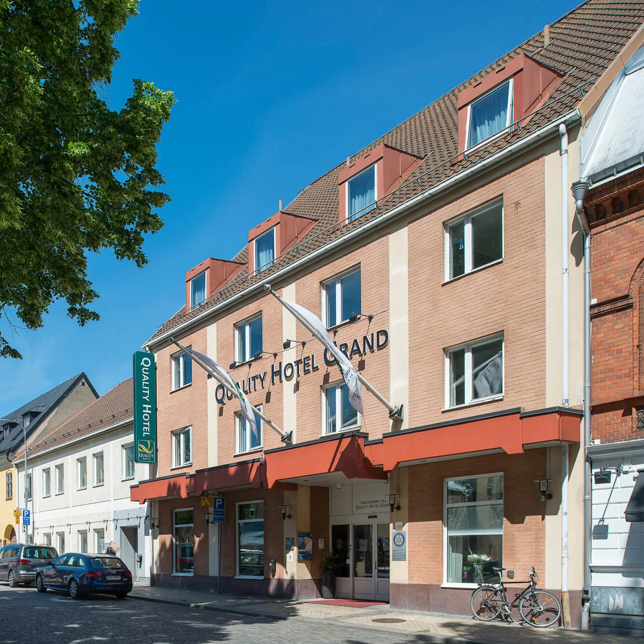 Quality Hotel Grand-byggnaden står på en kullerstensgata. Parkerade bilar kantar vägen. Gröna träd ger skugga. Text: "QUALITY HOTEL GRAND" och "Quality Hotel".