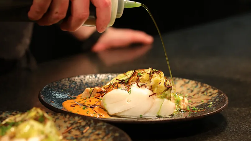 A hand drizzles green oil over elegantly plated fish with garnish on a dark ceramic plate, set against a smooth black countertop.