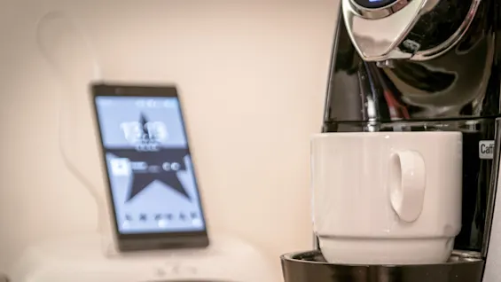 A coffee machine brews into a white cup; in the background, a smartphone displays "13:13 Fri," connected to a white dock.