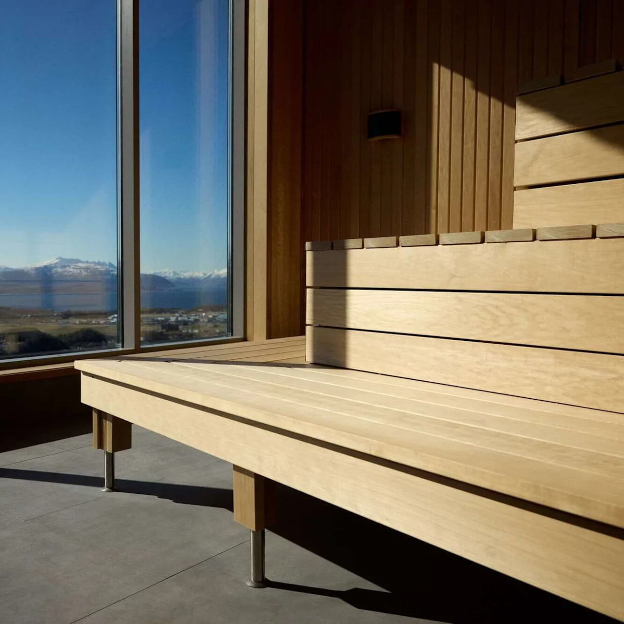 Wooden sauna bench bathed in sunlight, overlooking a vast landscape through large windows showing snowy mountains and a clear blue sky.