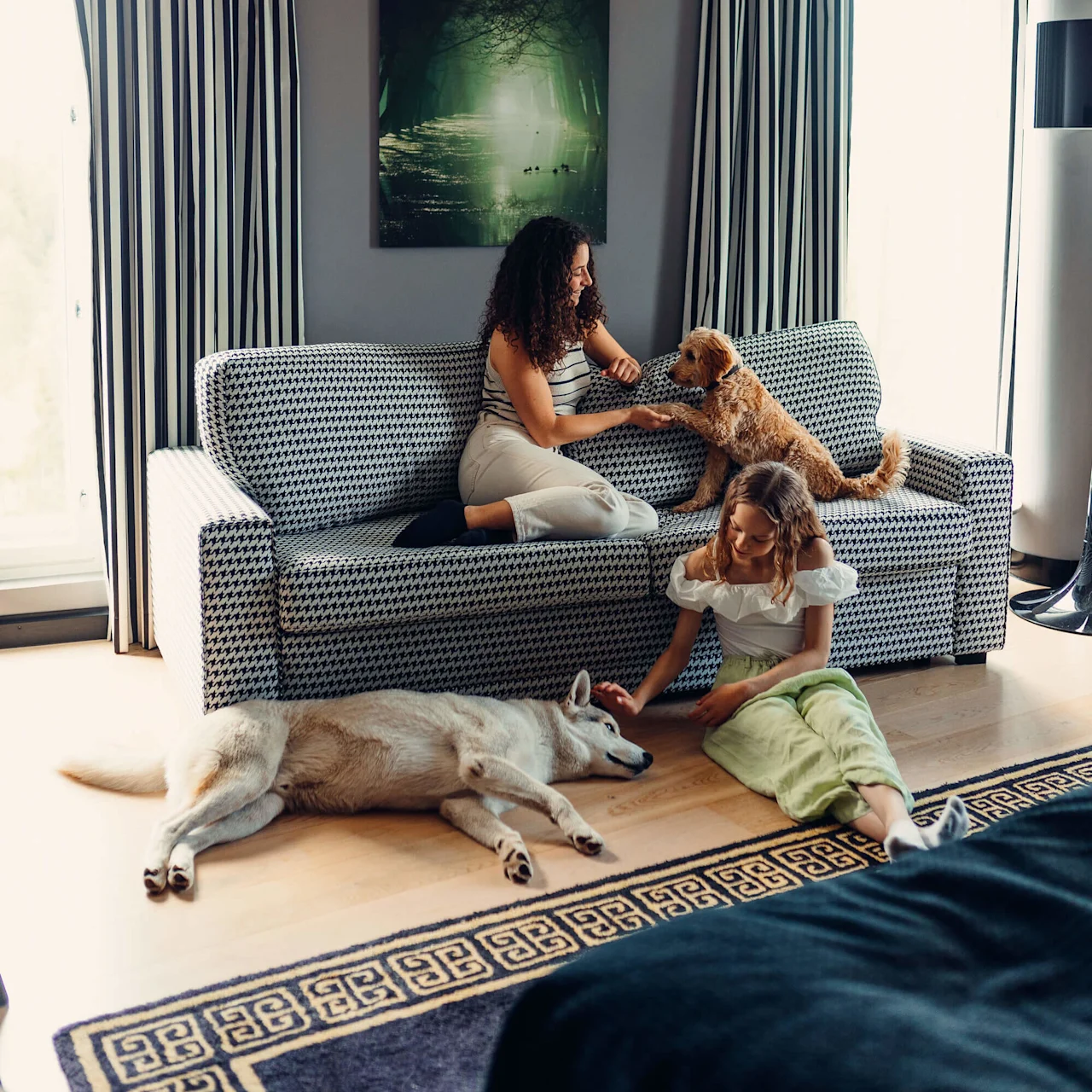 Two women interact with dogs; one sits on a patterned sofa playing with a brown dog, while the other sits on the floor petting a resting white dog. Softly lit room.