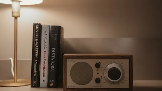 En mysig scen vid sängen med en lampa, böcker och en radio i vintagestil. Radion visar HOME HOTEL MODEL ONE SI.