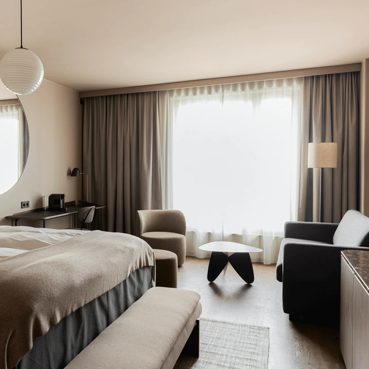 A modern hotel room features a neatly made bed with a round mirror, a small desk with a chair, and a seating area with an armchair, sofa, and table beside large curtained windows.