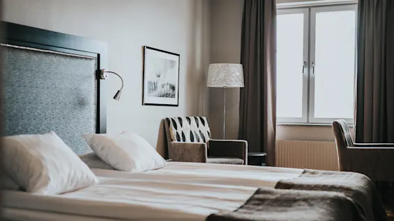 En pænt redt seng dominerer et varmt oplyst hotelværelse; en lænestol og en standerlampe tilbyder en hyggelig læsekrog ved vinduet.