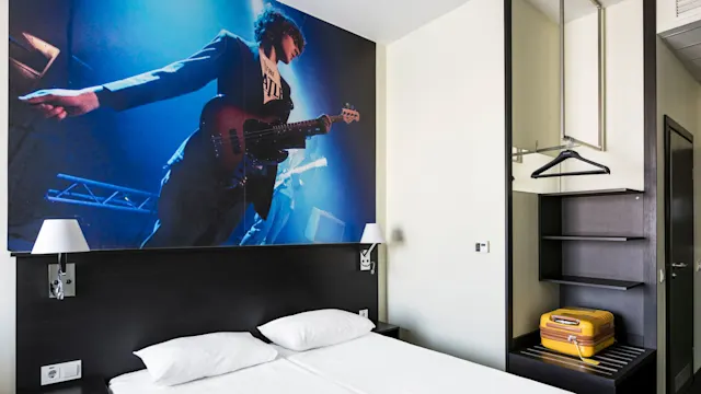 A hotel room features a double bed beneath a wall mural of a guitarist on stage. A wardrobe with shelves and a yellow suitcase is on the right.