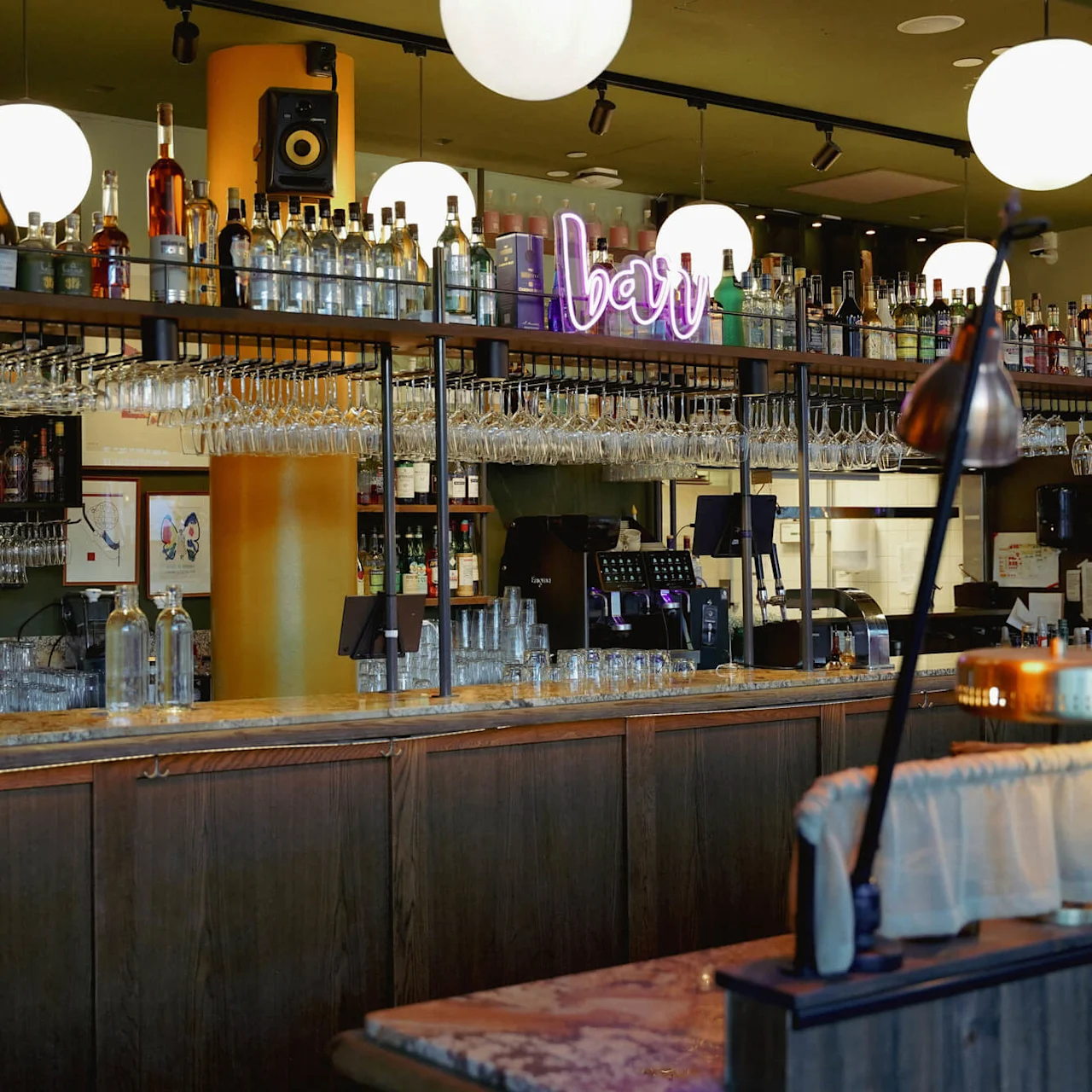 Træbardisk med glas og spiritusflasker samt et neonskilt der siger "bar" inden i en varmbelyst etablissement.