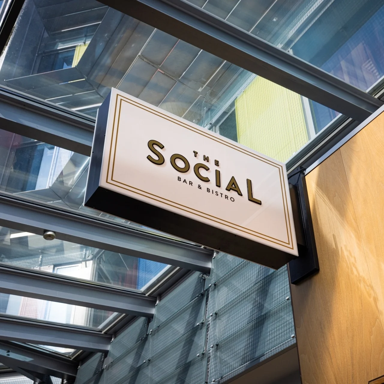 A sign for "THE SOCIAL BAR & BISTRO" hangs under a glass ceiling, angled towards a wooden wall, within a bright, modern architectural setting.