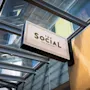 Et skilt for "THE SOCIAL BAR & BISTRO" henger under et glasstak, vinklet mot en trevegg i et lyst, moderne miljø.
