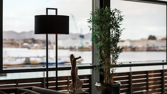 En svart gulvlampe står ved siden av en statue og en høy potteplante i en moderne stue med store vinduer som vender ut mot en vannkant og fjerne bygninger.