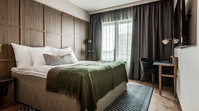 En seng med hvite puter og et grønt teppe står mot en trepanelvegg. Et skrivebord med stol og lampe er ved siden av, med gardiner og et vindu i bakgrunnen.