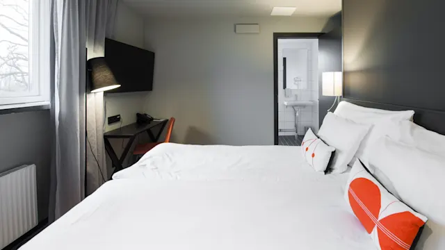 Siististi pedattu parisänky oranssein tyynyin on modernissa hotellihuoneessa. Työpöytä, tuoli ja lamppu ovat seinää vasten, ja ovi johtaa valoisaan kylpyhuoneeseen.