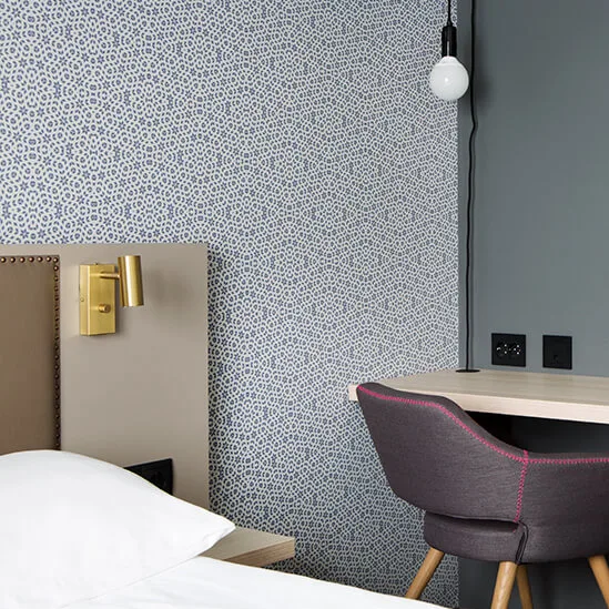 A modern desk with an adjustable lamp, positioned in a bedroom with patterned wallpaper and a bed partially visible.