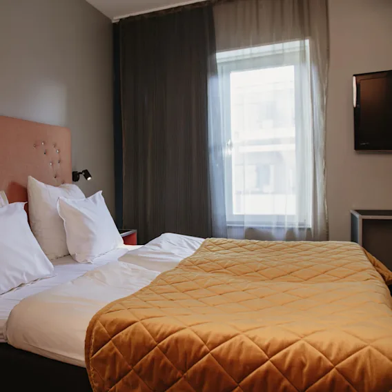 A bed with white pillows and an orange quilted blanket is in a hotel room. It has a tufted headboard, bedside tables, wall lamps, a window, and a TV.
