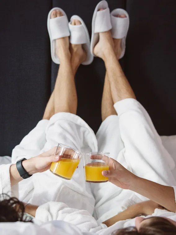 Två personer i vita morgonrockar ligger på en säng, håller glas med apelsinjuice och har fötterna i tofflor uppställda mot en mörk sänggavel.