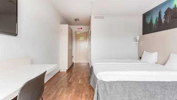 A modern hotel room with two beds, a desk, and a nature mural above the beds. An exit sign is visible near the door.