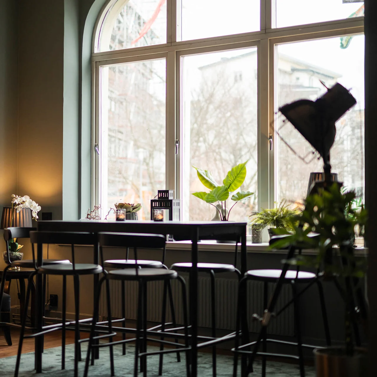 Ett högt bord med pallar i ett mysigt rum fyllt med växter, en golvlampa och stora fönster med utsikt över stadsbild, vilket skapar en inbjudande och avslappnad atmosfär.