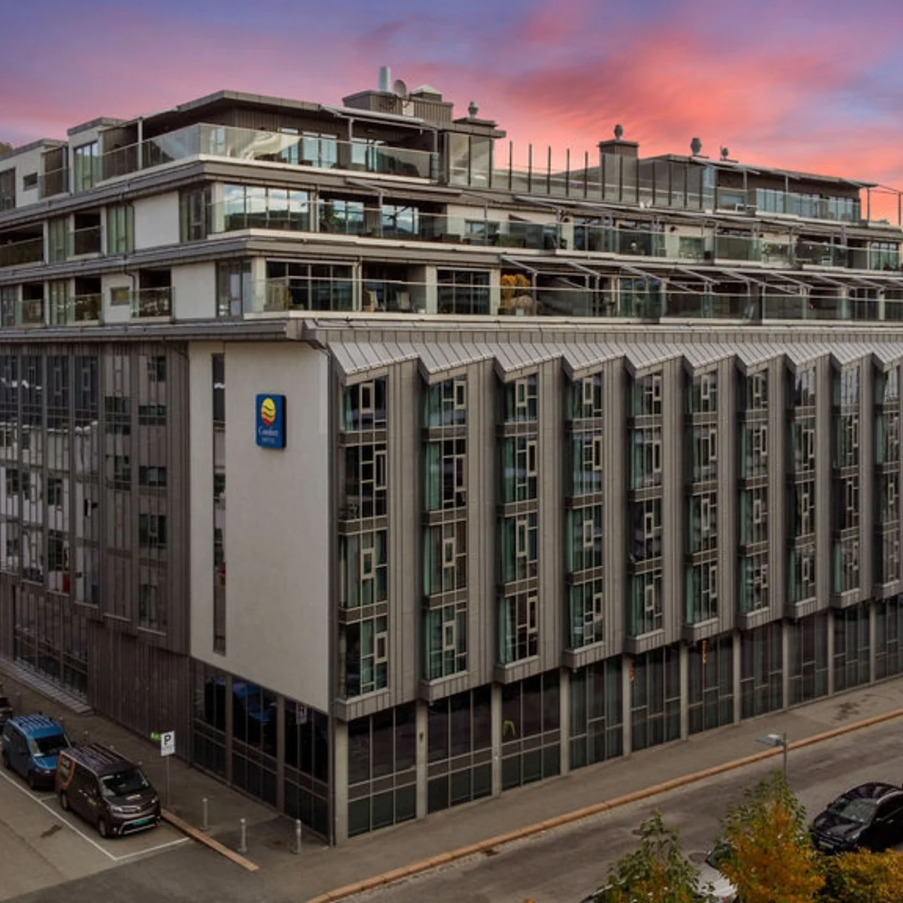 A multi-story building with geometric facade patterns stands in an urban area at dusk. The logo of Comfort Hotel is prominently displayed on the wall. Cars are parked nearby.