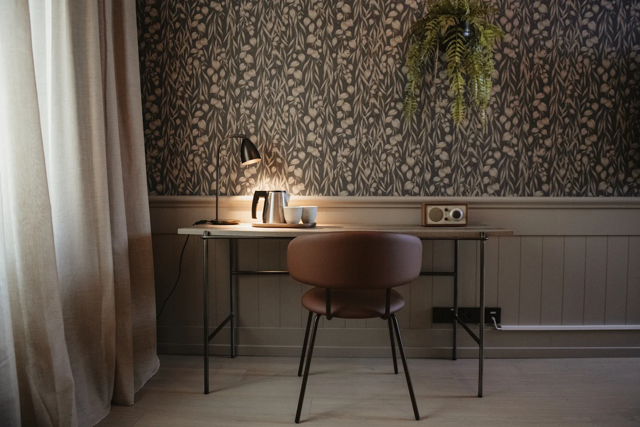 En funktionel og indbydende skrivebordsplads med en lampe, elkedel og radio, mod en baggrund af botanisk tapet.