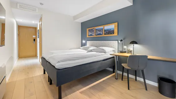 A neatly made bed in a clean, modern hotel room with wooden flooring, desk, chair, and decorative pictures on the wall.