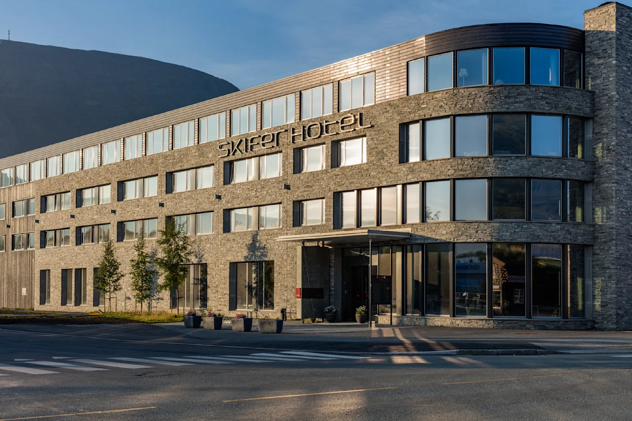 En moderne, fleretasjes bygning merket "Skiper Hotel" står ved en gate og reflekterer sollys. Bygningen har store vinduer og steinfasade i kupert terreng.