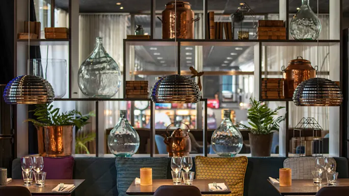 Tre bord med glass og servietter i en koselig restaurant, omgitt av dekorative glassflasker, kobberkjeler og planter, under metallpendellamper.