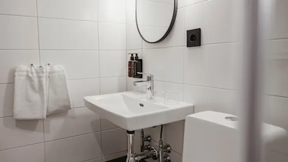 Ett rent, modernt badrum med vita kaklade väggar, ett handfat med toalettartiklar, en spegel och fräscha handdukar.