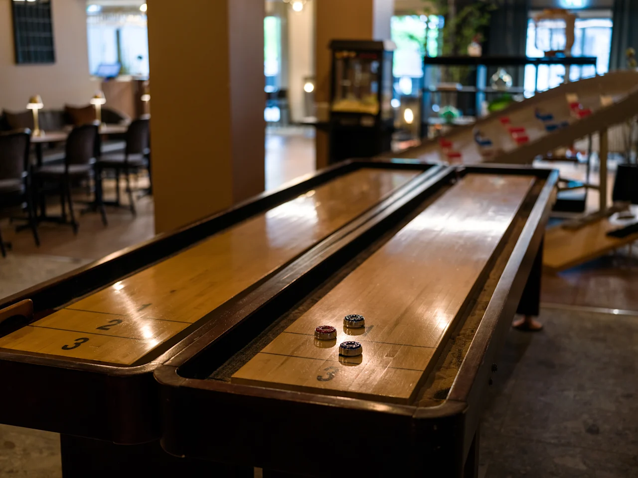 Ett shuffleboardbord med fyra puckar står i ett svagt upplyst rum omgivet av stolar och olika arkadspel.
