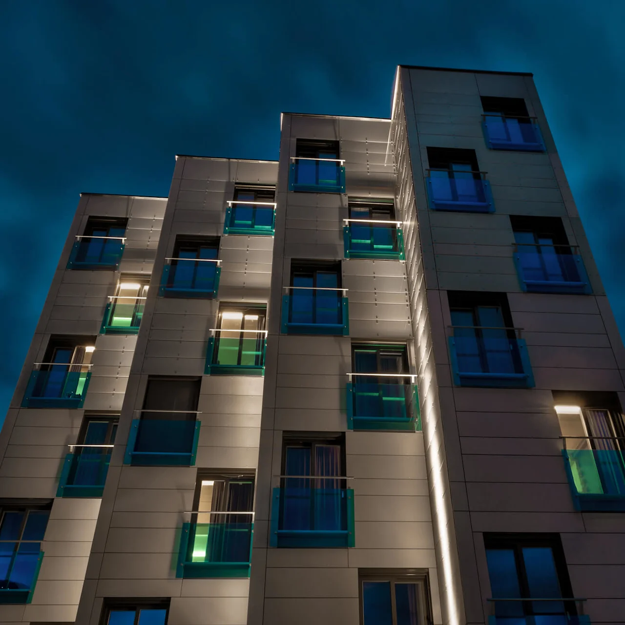 En modern lägenhetsbyggnad står upplyst på natten, med belysta fönster och reflekterande glasbalkonger mot en mörk, molnig himmel.