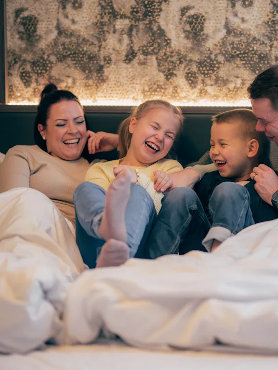 Nelihenkinen perhe pötköttelee hotellisängyssä valkoisen peiton alla nauraen yhdessä.